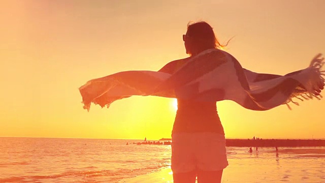 详情图-慢镜头:日落时分，戴着太阳镜和飘动的围巾的女孩在海滩上旋转