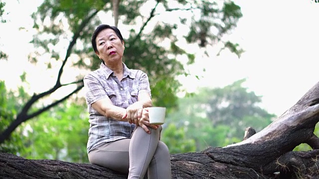 孤独的亚洲老年妇女坐在公园里独自喝咖啡，看起来很悲伤，思考着生活