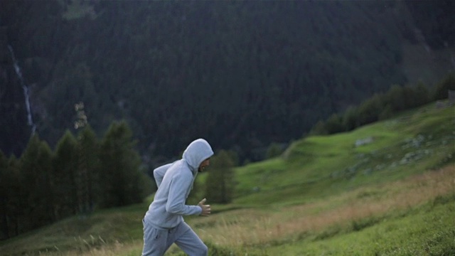 男人慢跑，跑上山，小山面对近距离的慢镜头。戴着兜帽的跑步者穿着灰色运动服运动鞋在草地上冲刺，努力训练马拉松。运动健康的生活方式