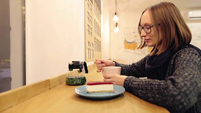 Сheerful女孩享受一杯热饮料和甜点坐在晚上在咖啡馆。迷人的白种女人戴着好心情的玻璃杯喝薄荷茶，肉桂，酸橙和蜂蜜