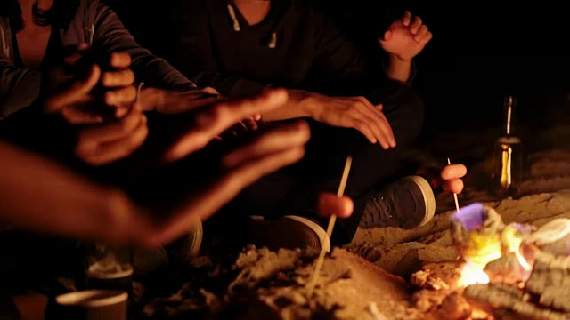 野餐的年轻人坐在篝火在海滩和温暖他们的手在晚上。特写视图