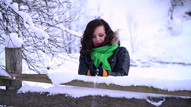 年轻美丽的女孩在白雪覆盖的公园里。她站在木栅栏附近。女孩把雪捧在手里，朝他吹了吹。高高兴兴地把雪堆起来。