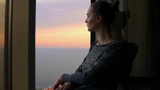 近距离观看美丽的年轻女子站在打开的窗户在日落的海边，享受的观点。背景是美丽的天空和大海