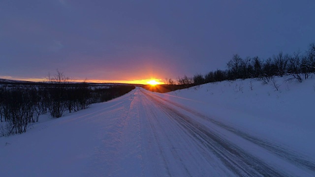 近距离观察:汽车在冰雪覆盖的森林道路上行驶在下雪的金色冬季日落