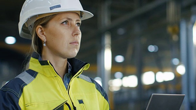 戴安全帽的女工业工程师站在重工业制造厂里使用笔记本电脑。在各种金属制品项目的背景零件躺着