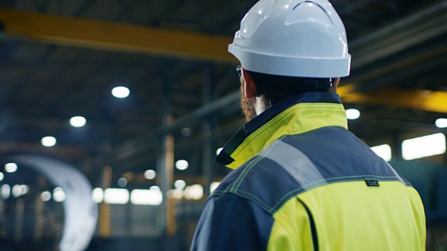 戴安全帽、穿安全服的工业工程师在大型重工业制造工厂四处查看。弧查看录像。
