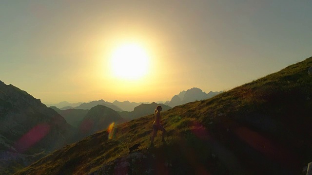 空中剪影:一名徒步旅行者和一只狗爬上长满草的小山，镜头从头顶飞过