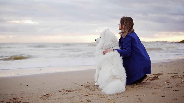 一个年轻女子坐在沙滩上，拥抱着她的萨摩耶犬在海边。白色毛茸茸的宠物在海滩上看着大海。真正的友谊。Slowmotion拍摄