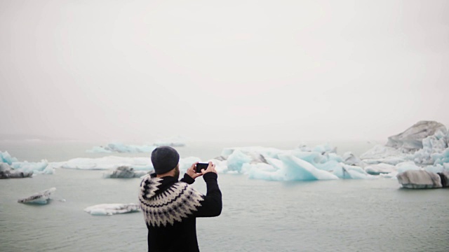 年轻的旅行男子站在冰岛的Jokulsalon冰礁湖，用智能手机拍摄冰川的背影