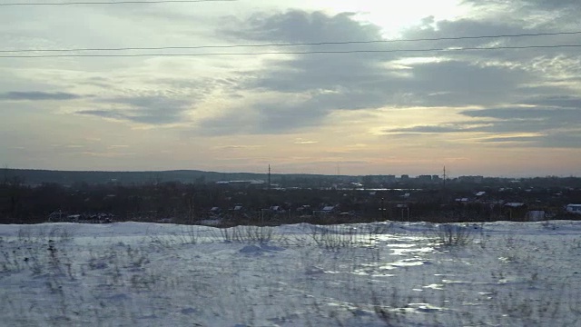 从移动的汽车或火车的窗口拍摄的冬季城市景观。镜头掠过雪堆、灌木丛、花园、避暑别墅和远处的一座城市。