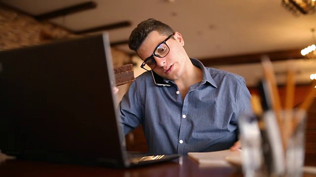 哈里英俊的年轻商人戴着眼镜在笔记本电脑上工作，打电话，做笔记，在笔记本上搜索信息，喝咖啡和吃蛋糕。多任务telecomuting概念