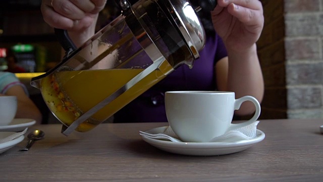 一个女人把沙棘茶从一个玻璃茶壶倒进一个白色的大杯子里
