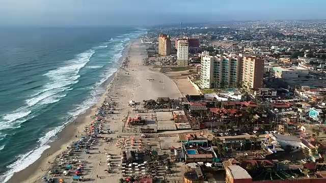 航拍向后飞行在繁忙的海滩，汹涌的海浪，和度假村在罗萨里托，墨西哥。