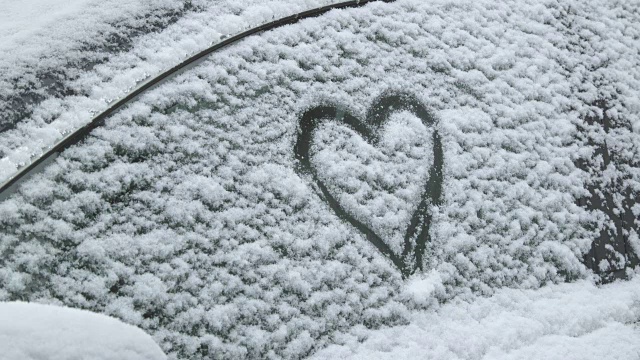 一辆被雪覆盖的汽车上的爱情告白——一个女人正在一辆被雪覆盖的汽车的侧窗上画一个心形