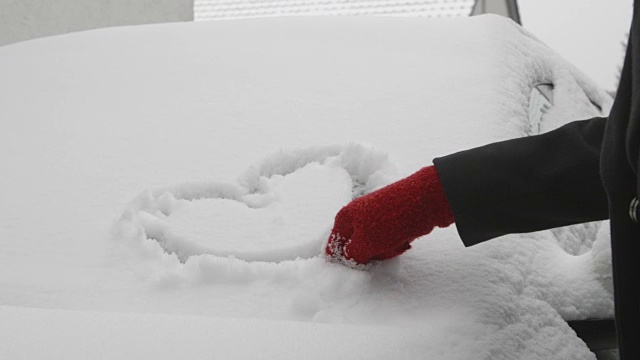 一辆被雪覆盖的汽车上的爱情告白——一个女人正在一辆被雪覆盖的汽车的挡风玻璃上画一个心形