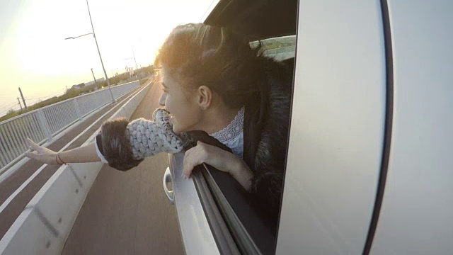 快乐迷人的女人在夕阳西下时，在汽车的后座上愉快地挥手，她的头伸出窗外