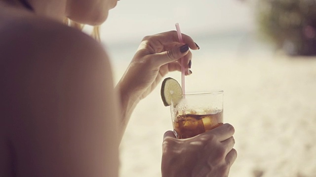 詳情圖-一個(gè)美麗的年輕女子在加勒比海炎熱的夏日的海灘上，用塑料吸管在一個(gè)裝飾有酸橙片的杯子里調(diào)雞尾酒