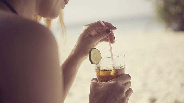 詳情圖-一個(gè)美麗的年輕女子在加勒比海炎熱的夏日的海灘上，用塑料吸管在一個(gè)裝飾有酸橙片的杯子里調(diào)雞尾酒
