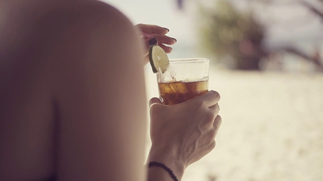 詳情圖-一個(gè)美麗的年輕女子在加勒比海炎熱的夏日的海灘上，用塑料吸管在一個(gè)裝飾有酸橙片的杯子里調(diào)雞尾酒