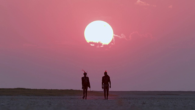 博茨瓦纳，两名圣布须曼人正穿过Makgadikgadi pan，身后是夕阳西下的红色天空