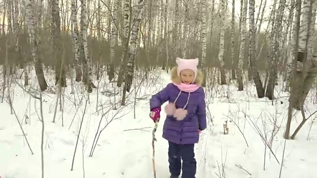 小女孩拿着一根棍子在白桦林里奔跑。年轻女孩开心的笑话和落在冬天的森林雪。