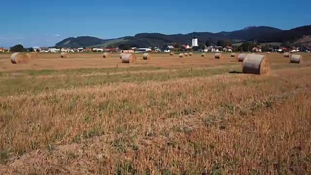 在有圆草堆的田野上空低空飞行。风景如画的小村庄和山峦为背景。