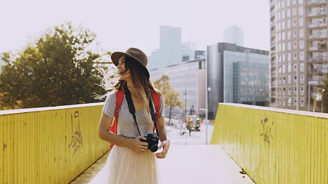 戴帽子，长发的女孩兴奋地走在桥上。欧洲女旅人开心，在黄墙之间转身。4 k