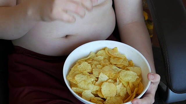 胖男孩在吃薯片。孩子腹部脂肪褶皱。不健康食品，快餐
