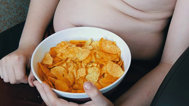身体上有脂肪褶皱的胖少年正在吃薯片。不健康食品，快餐。孩子的营养问题