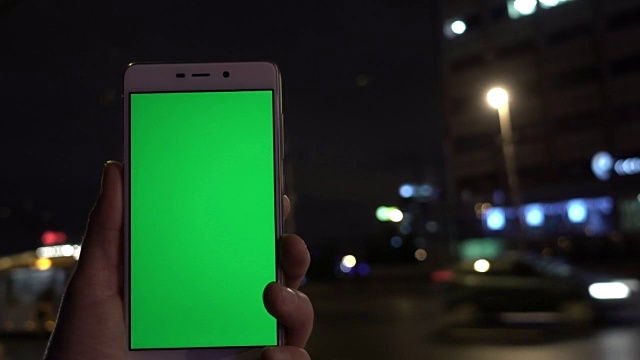 男子手握白色智能手机，在夜城灯光散景的背景下。绿色的屏幕。