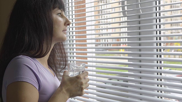 一位年轻女子正站在窗边喝着干净的淡水。女孩喝了一杯水，看着窗外。黑发的女人。