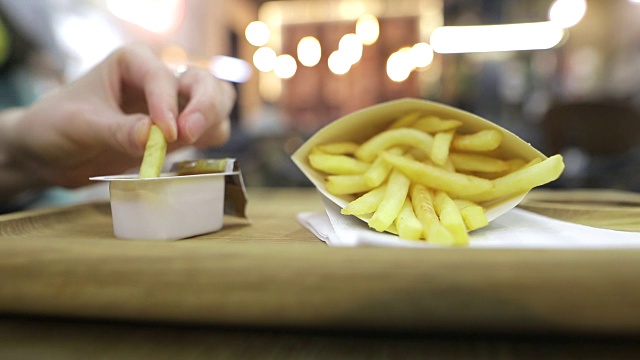 一个饥饿的人的手正在挑选快餐薯条，并把它们浸在甜酸酱。游客到咖啡馆和许多爱迪生灯在一个模糊的背景。晚上的时间