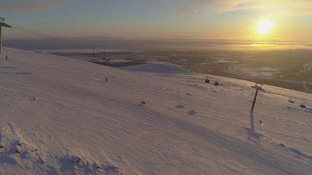 在阳光明媚的冬季日落，人们乘坐缆车在滑雪坡上滑雪和滑板。太阳在坐标系。鸟瞰图。无人机正在横向飞行。远景。