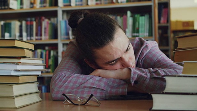 英俊的年轻男子坐在他的头在桌子上睡觉，在学校图书馆周围的大量书籍。他的眼镜放在他面前