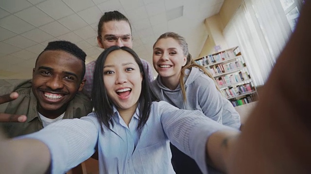 亚洲女孩拿着智能手机和开朗的同学自拍，在大学图书馆玩得开心