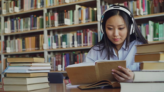 相当积极的亚洲青少年学生戴着耳机听音乐坐在大图书馆的桌子上拿着书和写总结