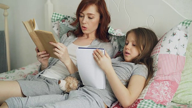 细心的妈妈帮她可爱的小女儿做小学作业。爱的妈妈在看书，女孩在抄写笔记，坐在家里的床上
