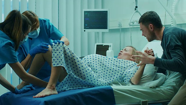 在医院里，妇女在分娩，产科医生和医生协助，她的丈夫通过牵手支持她。配备专业助产士的现代产科病房。