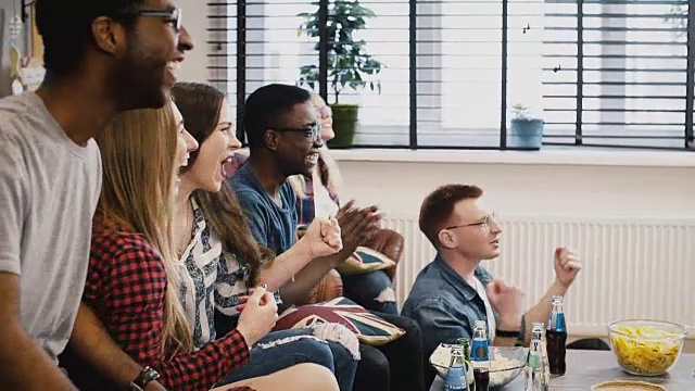 多民族的年轻人在电视上看足球。庆祝成功。球迷支持最喜欢的球队。零食和饮料