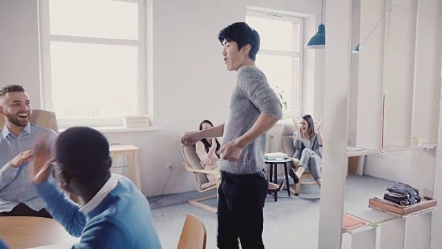 快乐的亚洲经理跳着非常有趣的庆祝舞走在现代的办公室里，多民族的同事鼓掌大笑4K