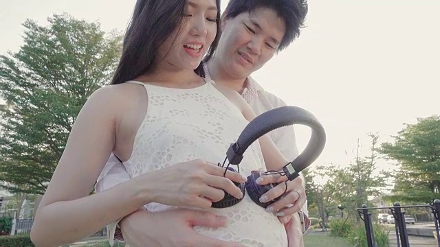 快乐怀孕的亚洲妇女和丈夫与耳机在她的肚子在花园。4 k