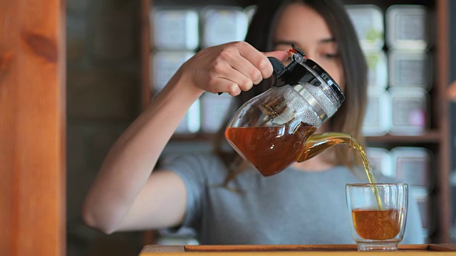 年轻微笑的女人从玻璃壶倒红茶到小杯子