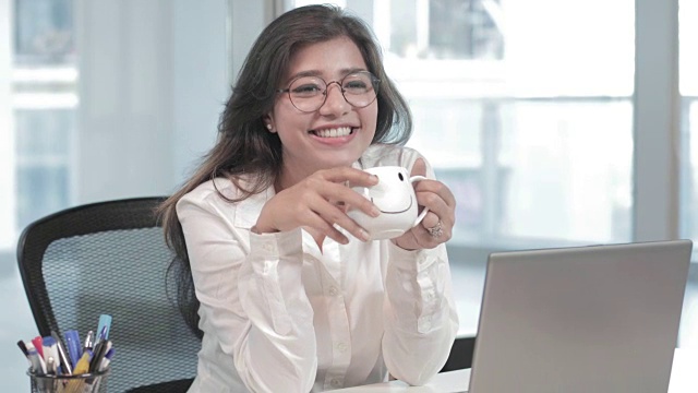 一位年轻漂亮的女职员坐在笔记本电脑前，喝着咖啡，面带微笑地坐在办公桌前。
