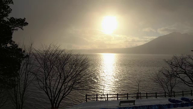 日出在波浪起伏的湖面上，冬天下雪，树木和长凳的剪影