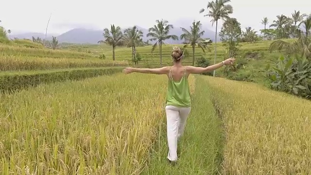 跟踪拍摄，年轻女子走在稻田里，周围是令人惊叹的绿色葱郁的自然。伸开手臂，慢动作投篮