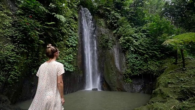 慢动作年轻女子享受新鲜瀑布在巴厘岛，印度尼西亚，亚洲。人们旅游乐趣自然的概念。女孩伸着胳膊