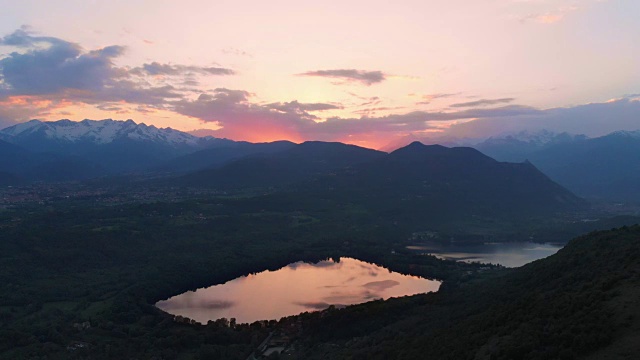空中:黄昏时分，意大利皮埃蒙特阿尔卑斯山脉上的乡村、森林林地和湖泊上空，日落时五彩缤纷的天空