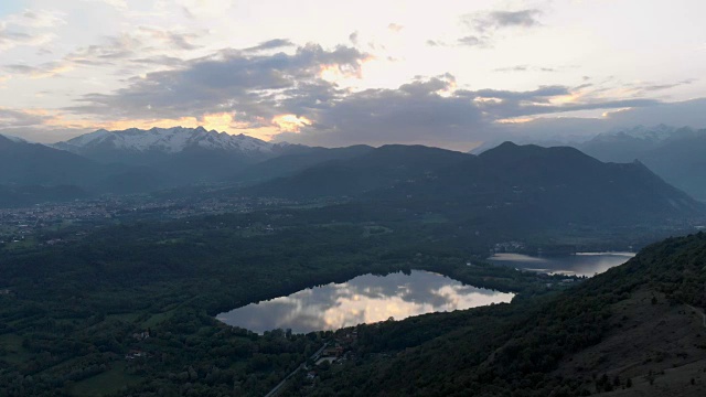 空中:黄昏时分，意大利皮埃蒙特阿尔卑斯山脉上的乡村、森林林地和湖泊上空，日落时五彩缤纷的天空