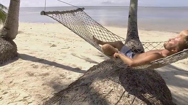年轻男子在菲律宾热带岛屿海滩的吊床上放松的慢动作视频。人们旅游度假放松的概念。