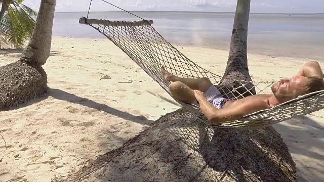 年轻男子在菲律宾热带岛屿海滩的吊床上放松的慢动作视频。人们旅游度假放松的概念。